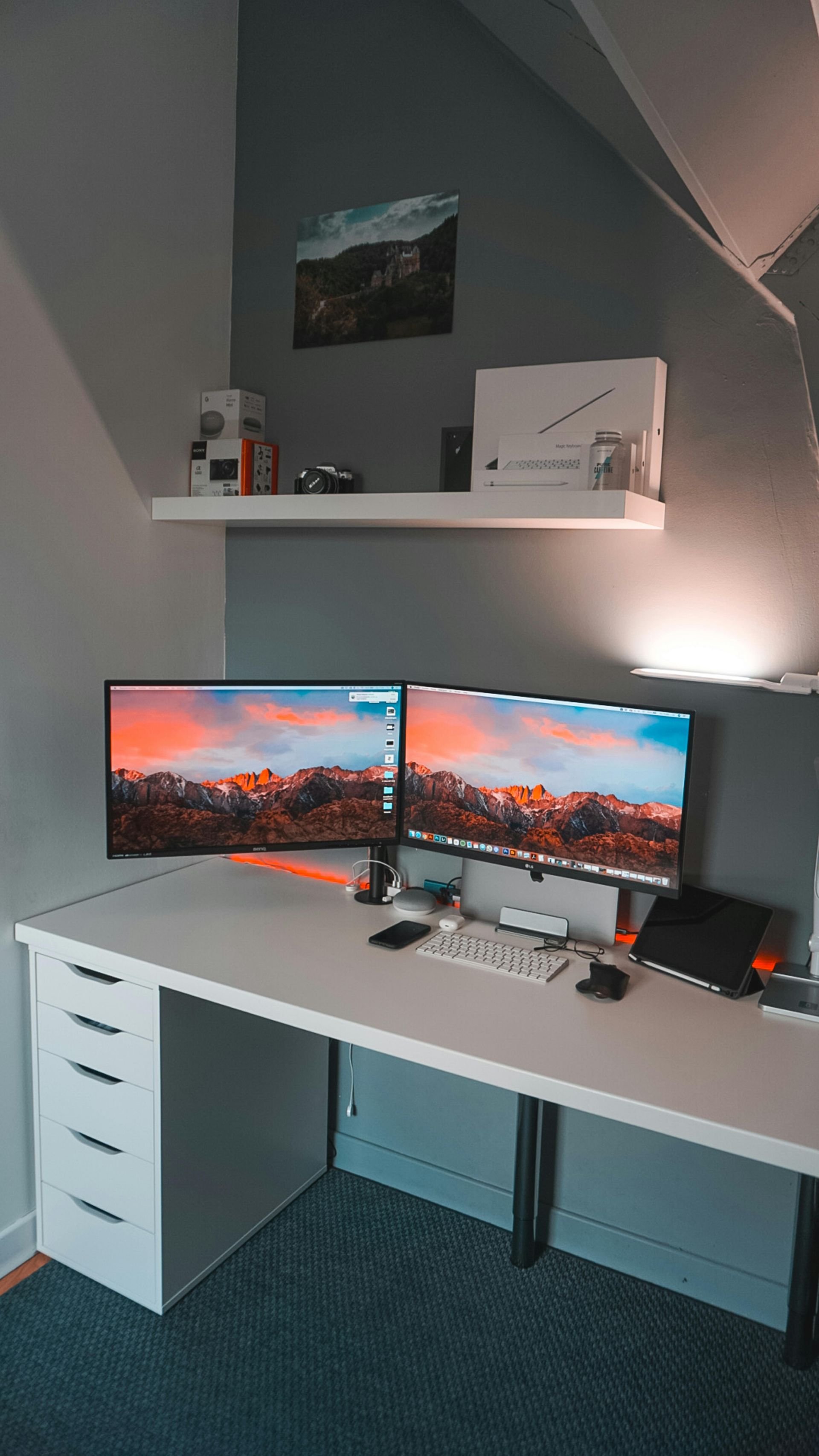 A dual-monitor desk setup in a room with a white drawer unit, a floating wall shelf, and a dark rug on the floor.