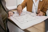 Two professionals in business attire discuss a floor plan document on a wooden desk.