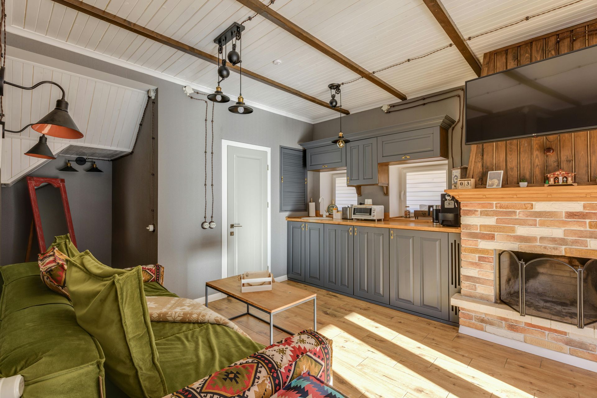 A rustic room with a green sofa, a coffee table, a grey kitchenette, and a brick fireplace.