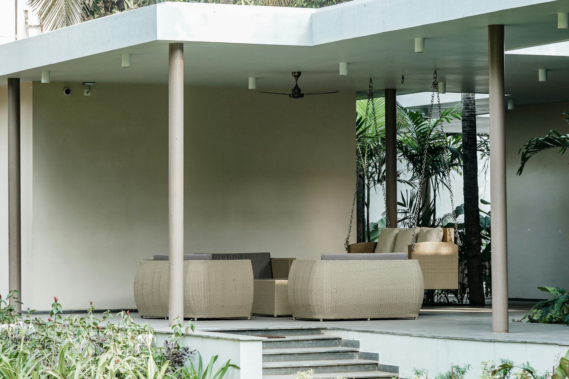 A covered outdoor patio with light-colored, woven lounge furniture and steps leading up from a garden area.