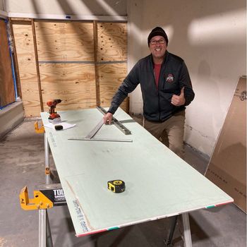 A man is standing next to a large piece of drywall and giving a thumbs up.