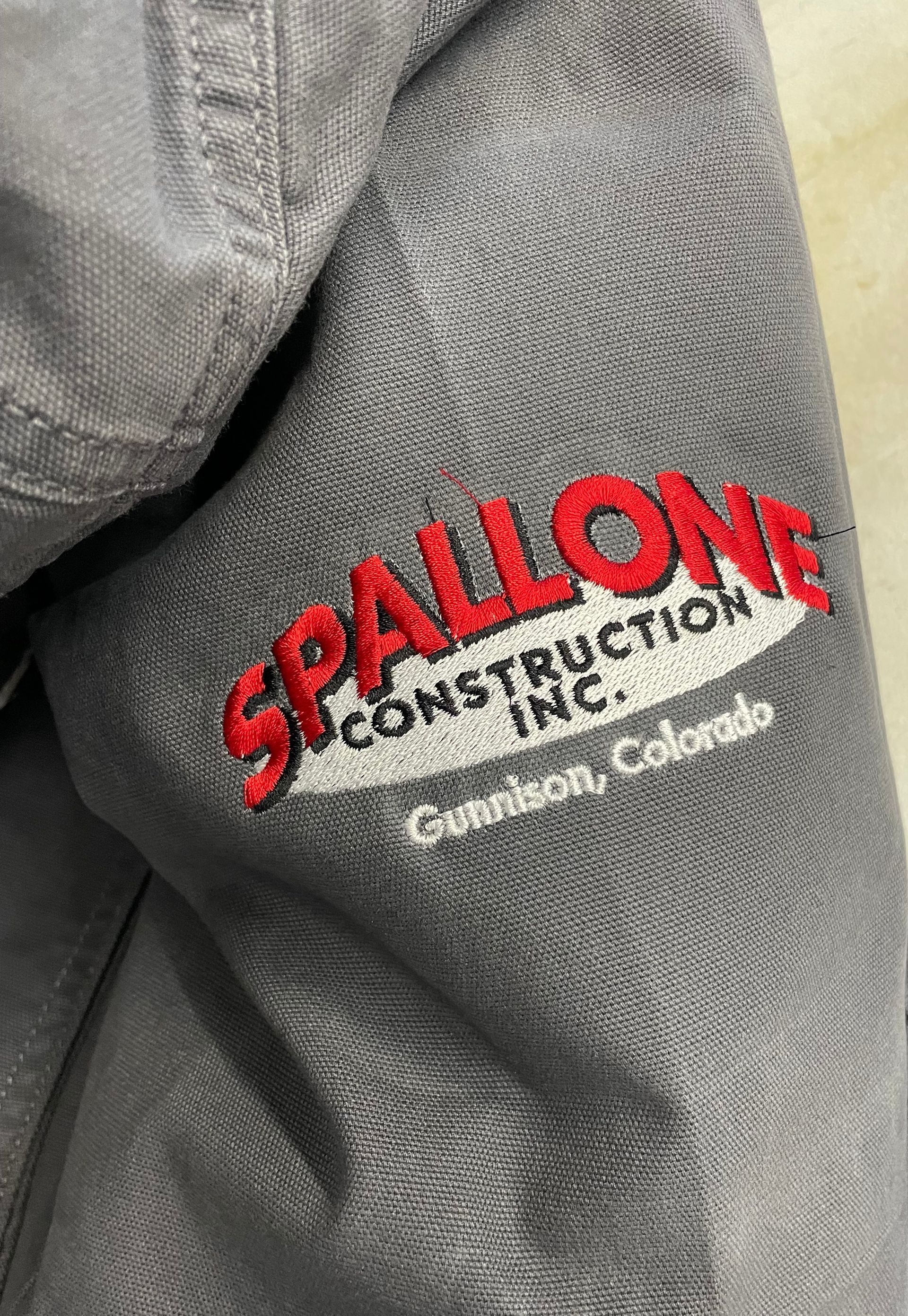 A person is wearing a gray jacket with a red embroidered logo on the sleeve.