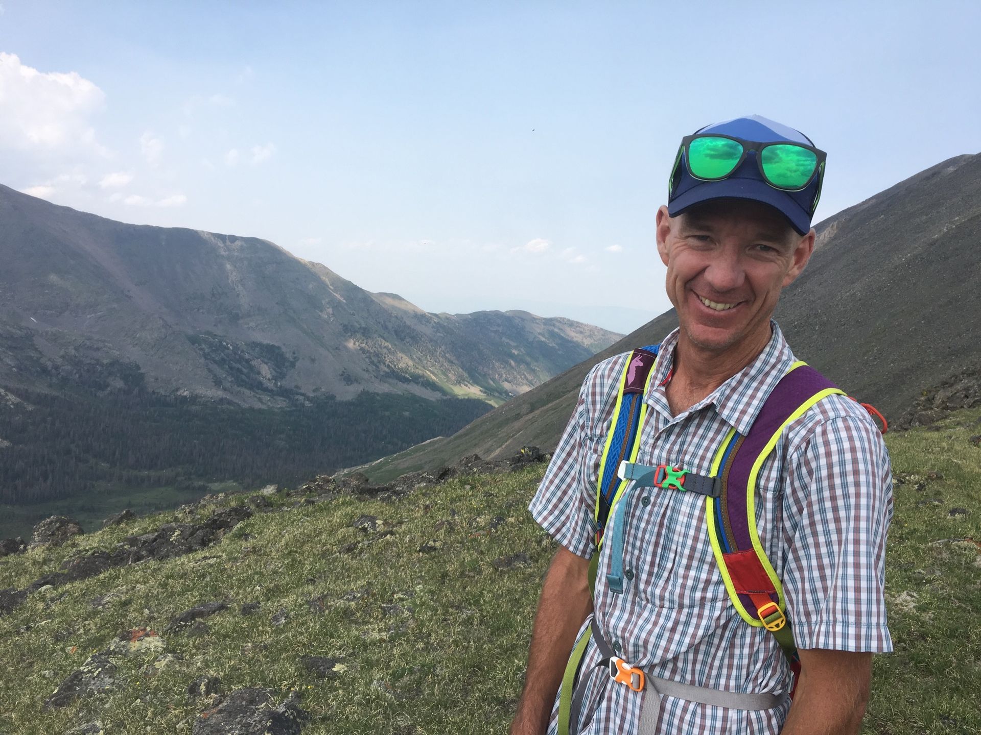 A man wearing sunglasses and a hat is standing on top of a mountain.