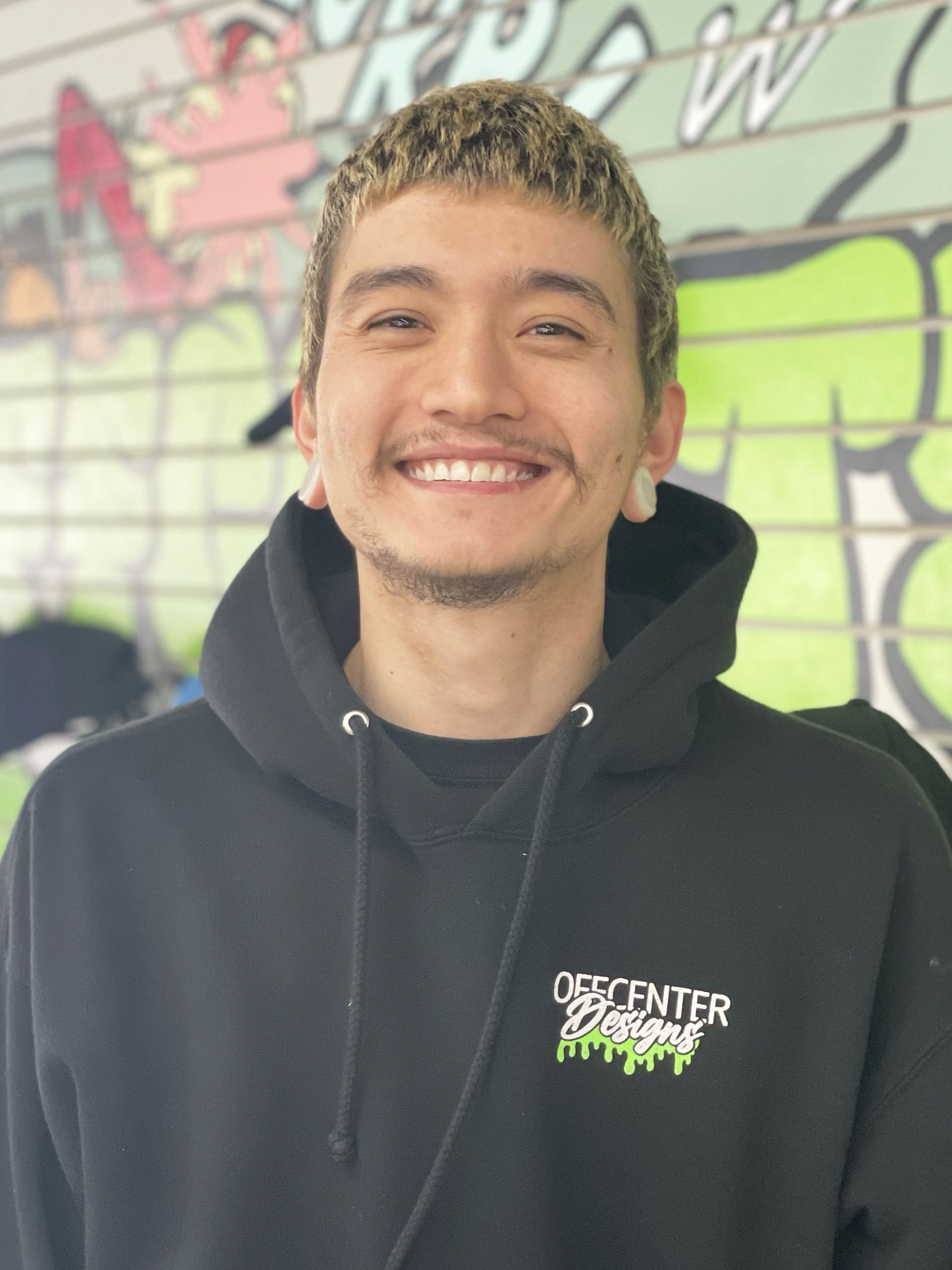 A man wearing a black hoodie is smiling in front of a graffiti wall.