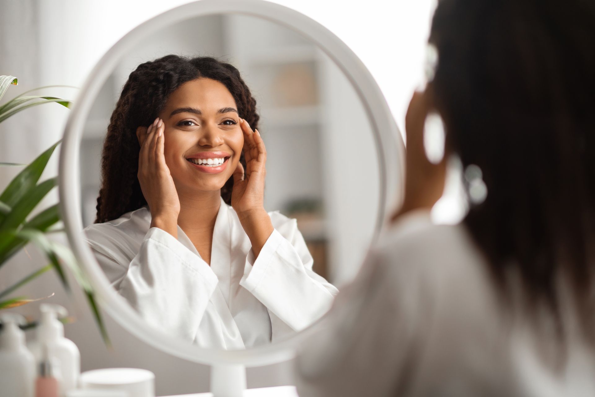Woman in a white robe smiles, touching her face while looking in a round mirror.