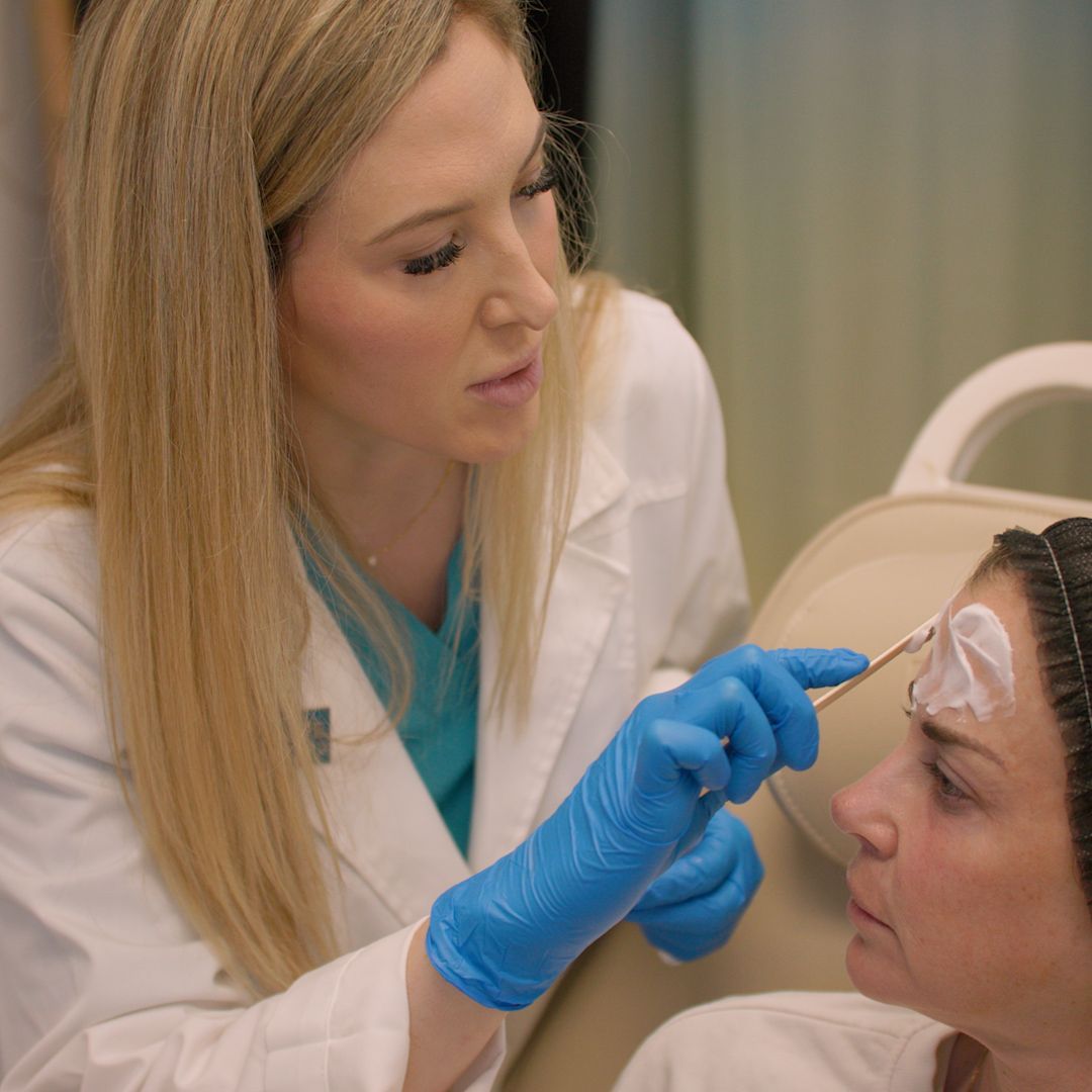 A woman in a white coat and blue gloves is applying cream to a woman 's forehead.