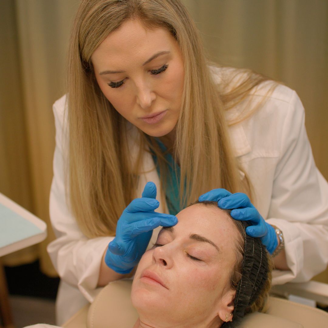 A woman in a white coat and blue gloves is examining another woman 's face.