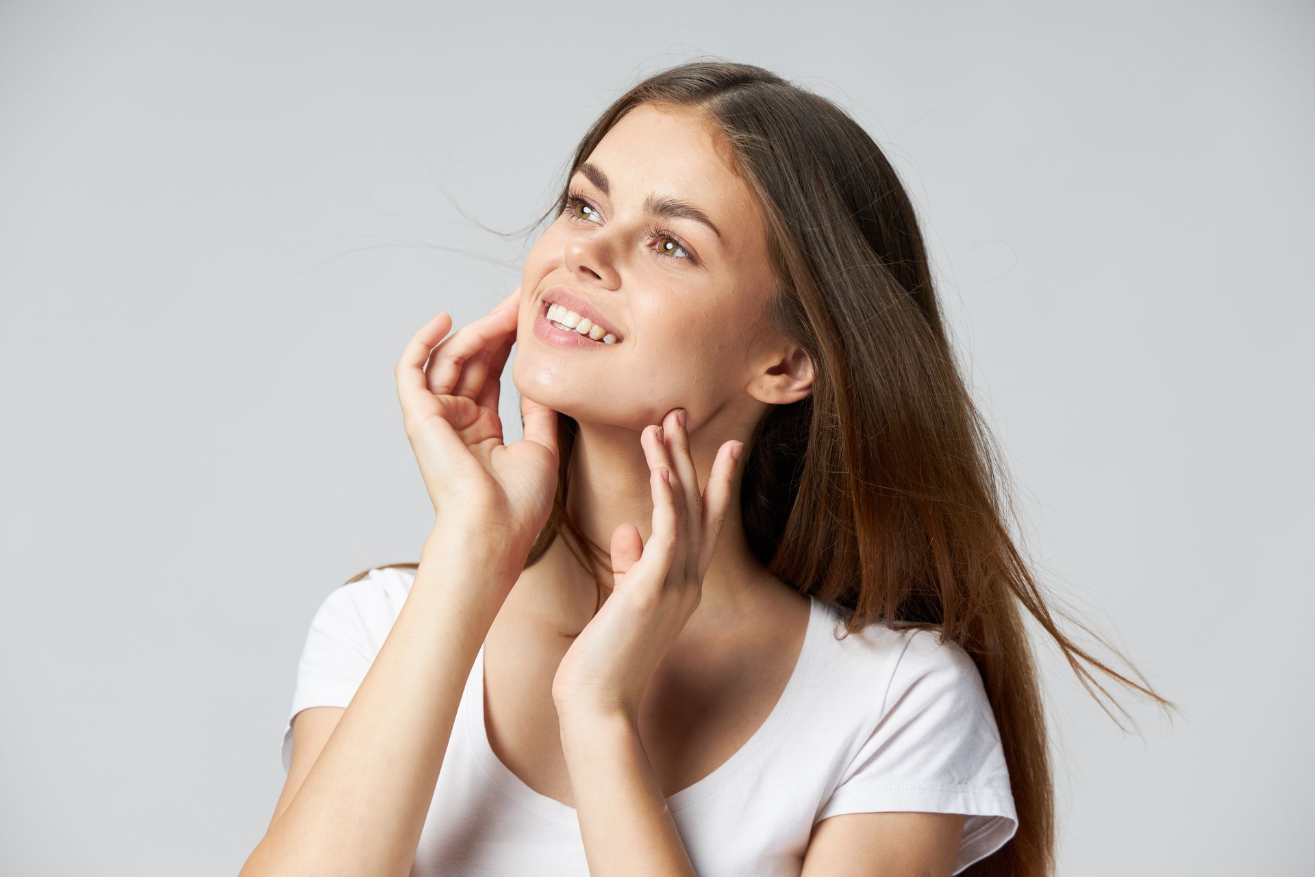 Woman with long brown hair, smiling, touching face, looking upwards.