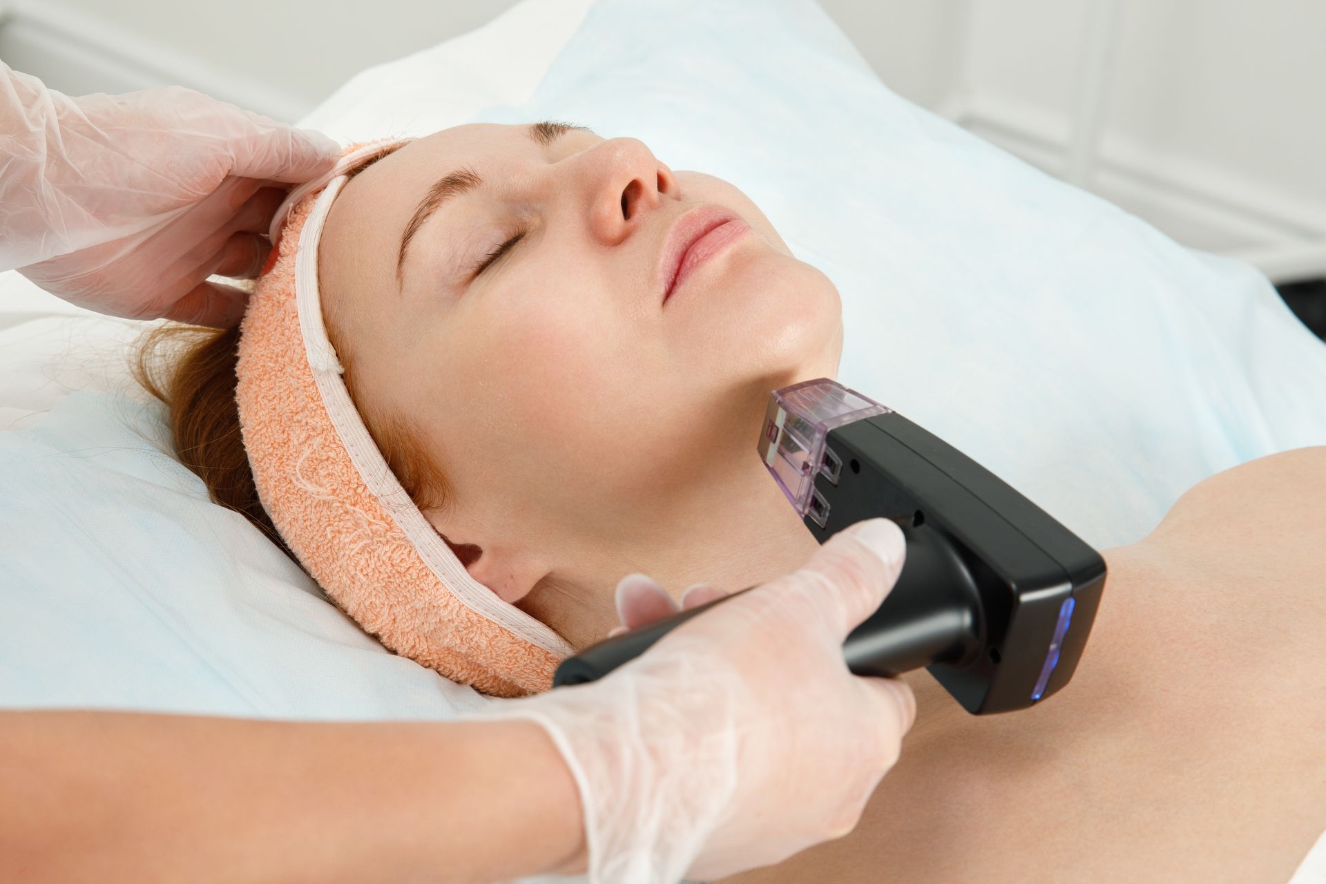 Woman receiving facial treatment, lying on a table. Hands with gloves holding a device on her face.