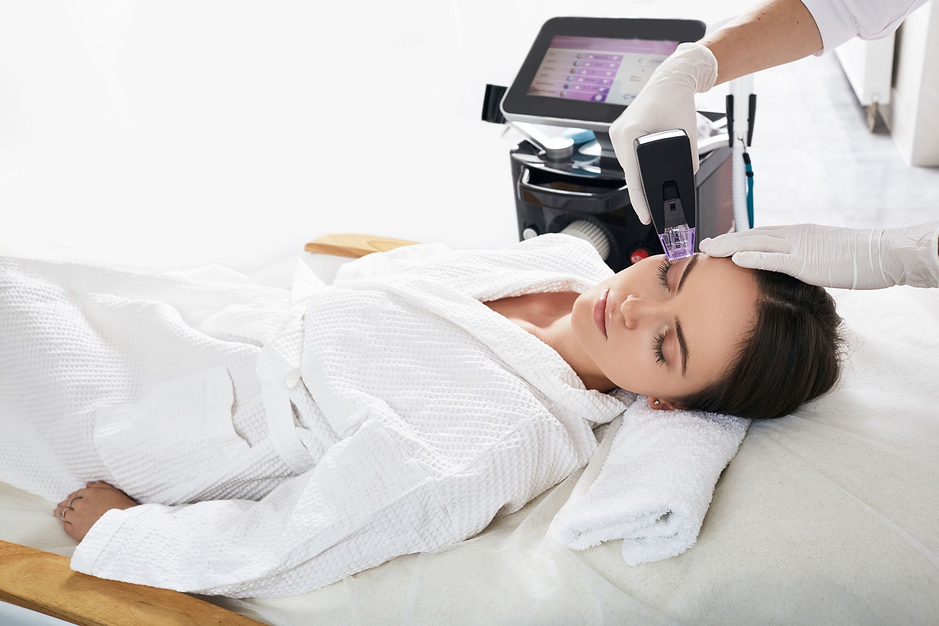 Woman receiving facial treatment with a medical device. She's lying down, eyes closed, and wearing a white robe.