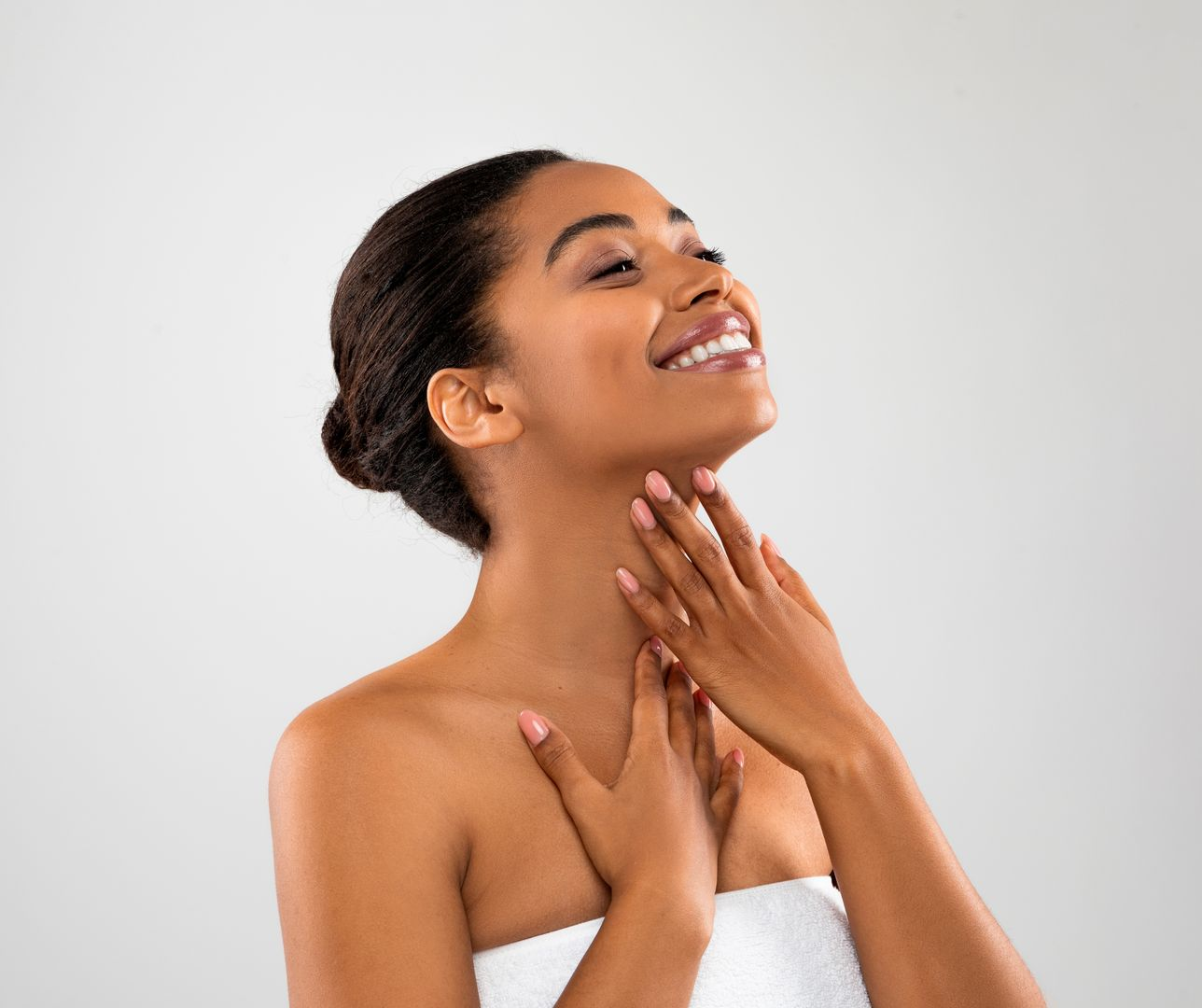 Woman touching her neck and smiling, wrapped in a white towel, against a gray background.