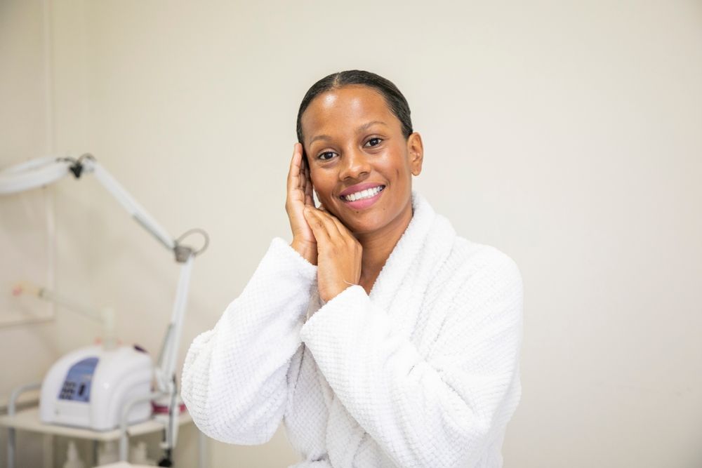 Woman in a white robe smiles, hands near face in a spa setting with equipment.