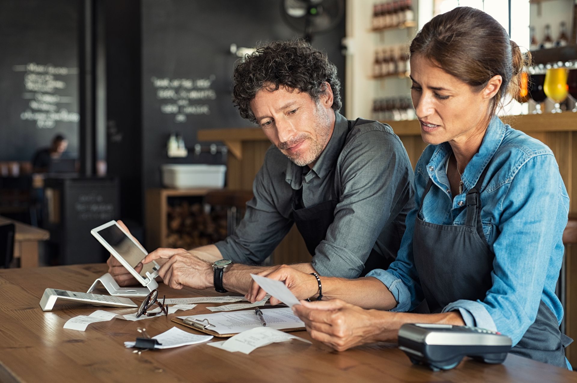 Two restaurant owners calculating receipts with a tablet and paperwork on a table.