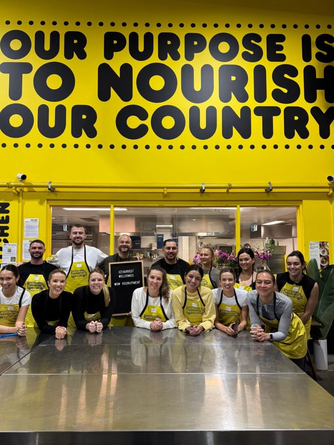 Group of people in yellow aprons and shirts, standing behind a stainless steel counter in a yellow kitchen.