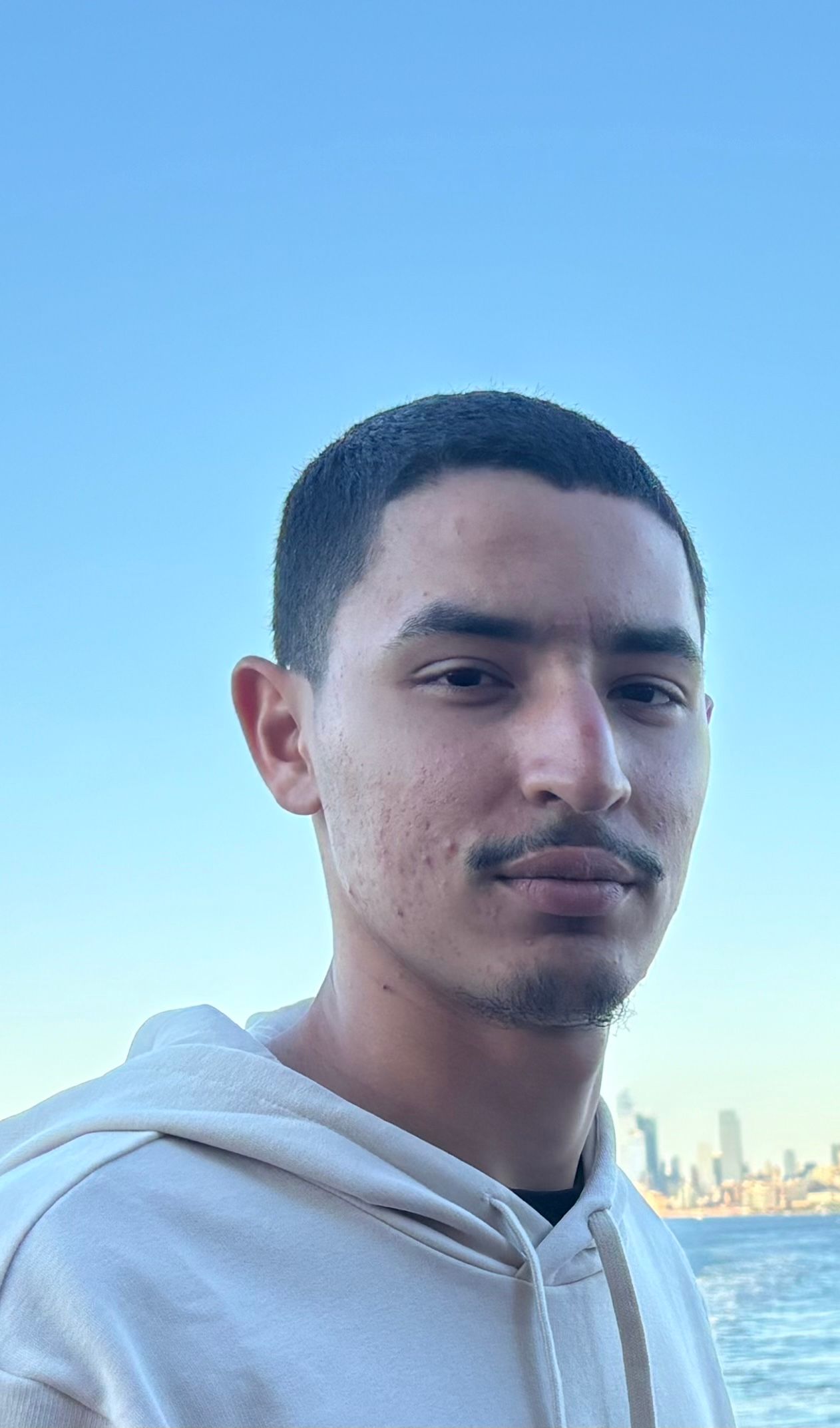 A young man in a white hoodie is standing in front of a body of water.