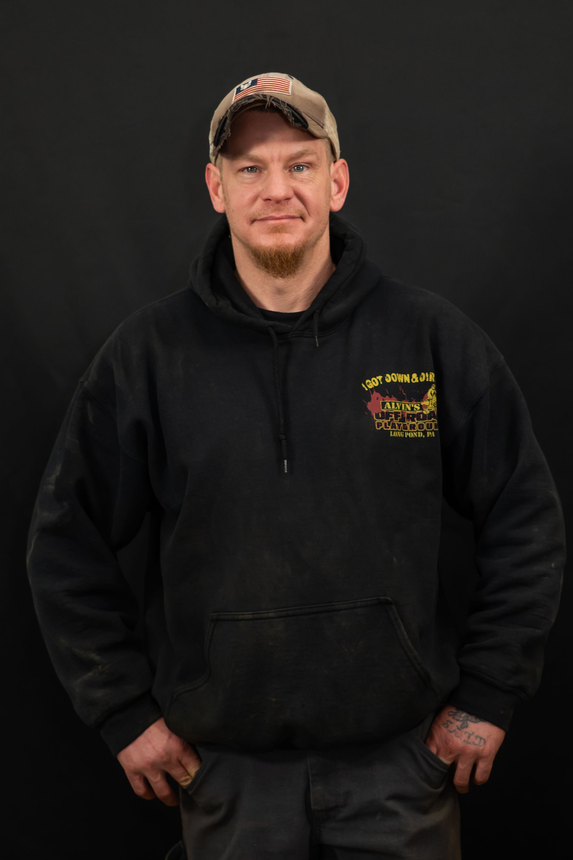 A man wearing a black hoodie and a hat is standing with his hands in his pockets.