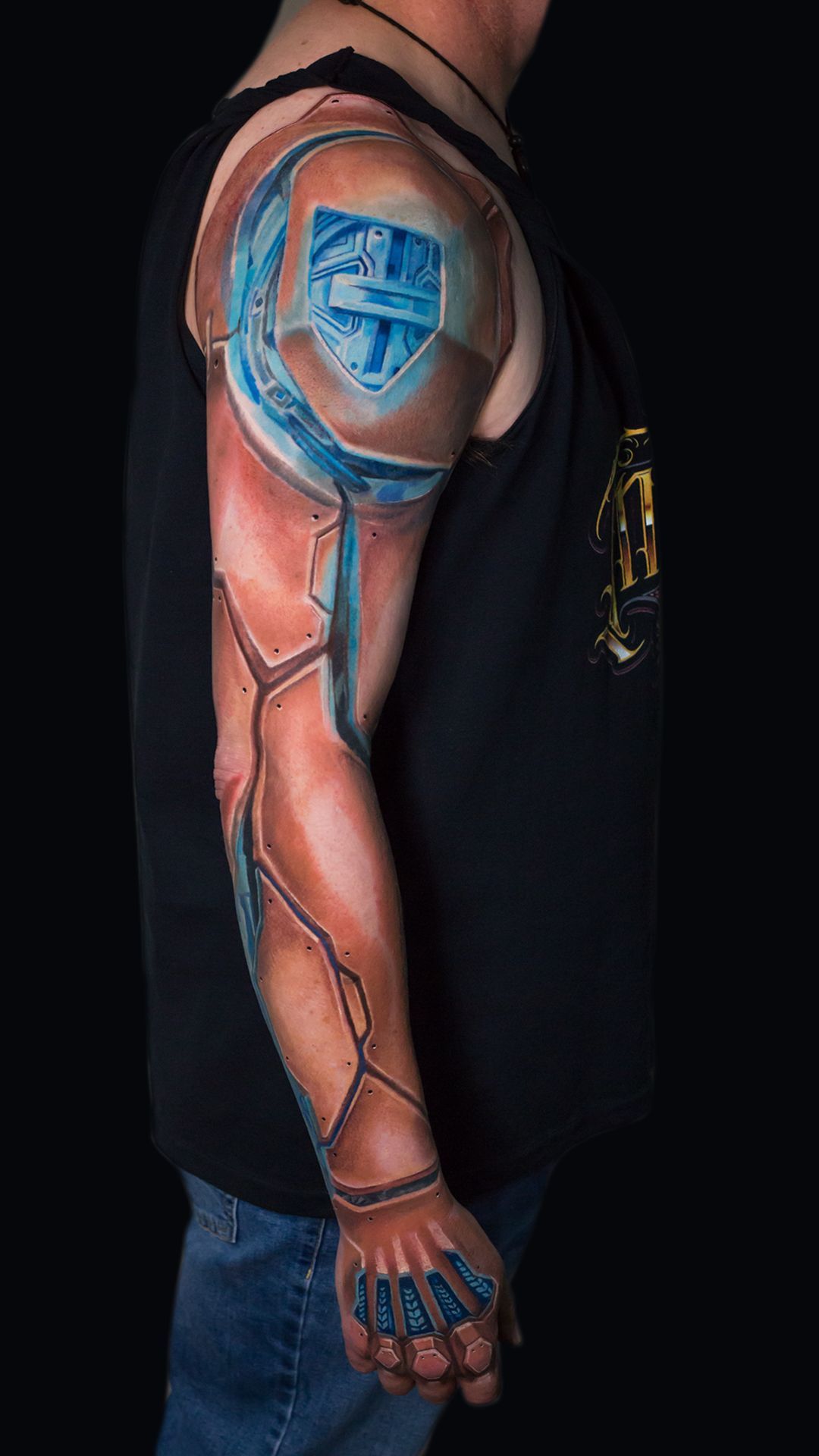 Side view of a tattooed arm with a blue shield tattoo, wearing a black sleeveless shirt and jeans.