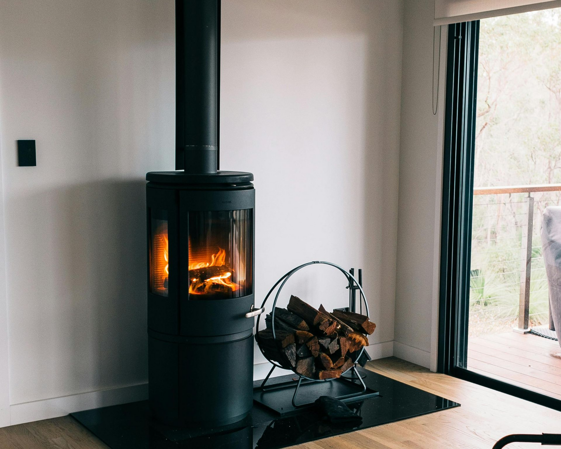 Poêle à bois noir avec un feu allumé, à côté d'un porte-bûches sur un plancher en bois, à côté d'une porte coulissante en verre.