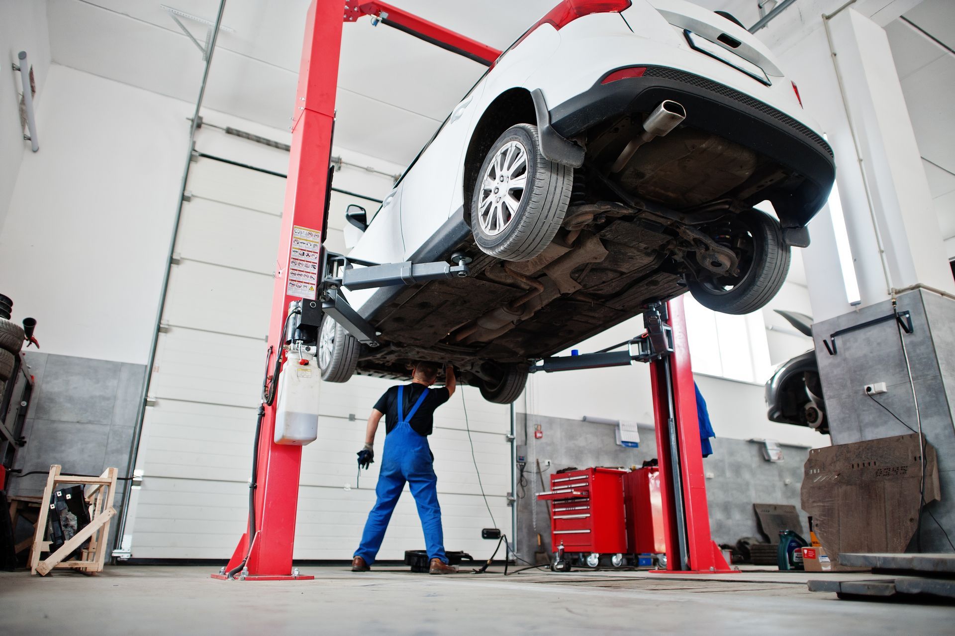 A worker in blue overalls inspects the underside of a white car raised on a red hydraulic lift in an auto repair shop.