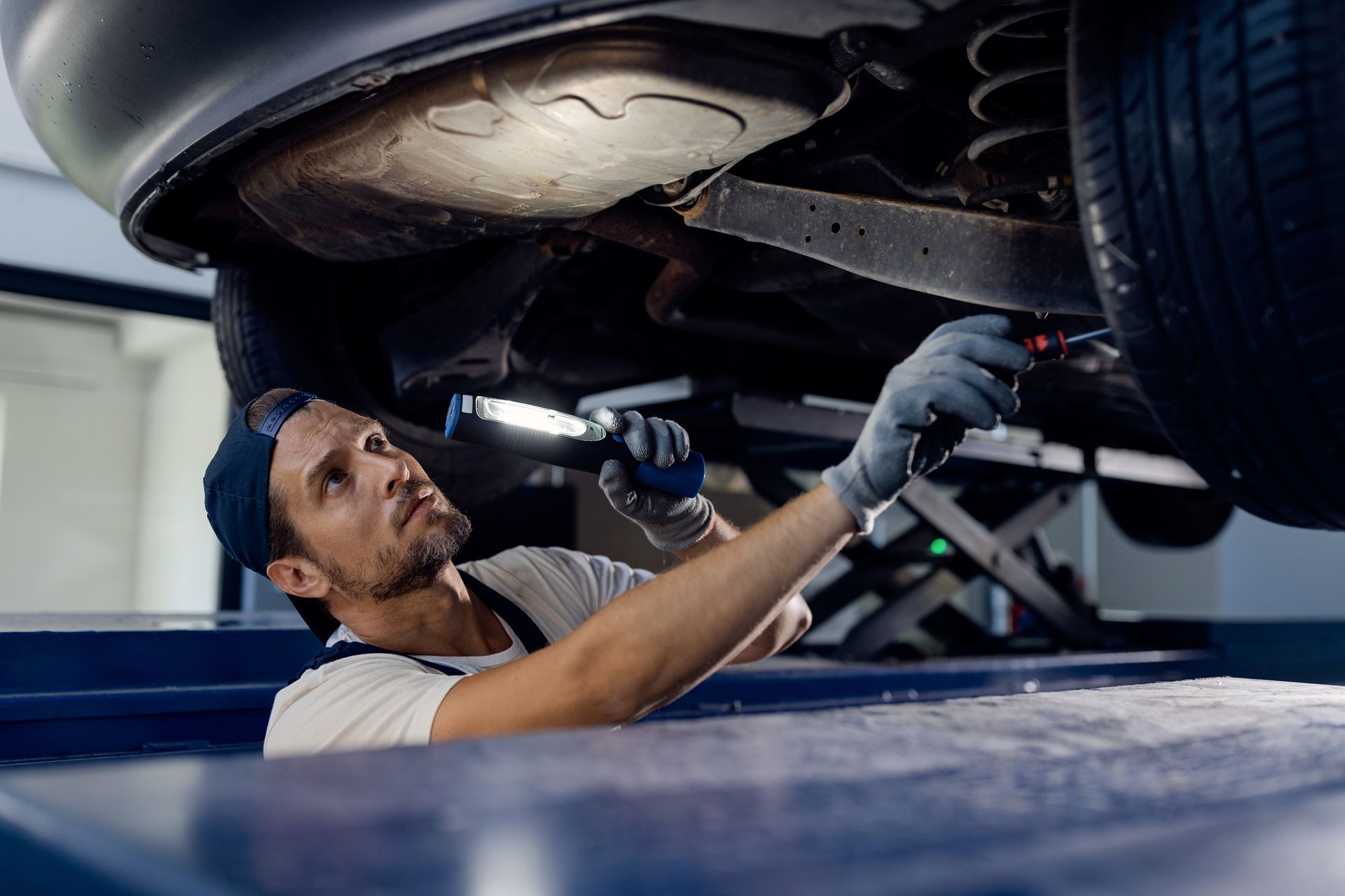A mechanic wearing gloves and a cap uses a flashlight to inspect the undercarriage of a car raised on a lift.