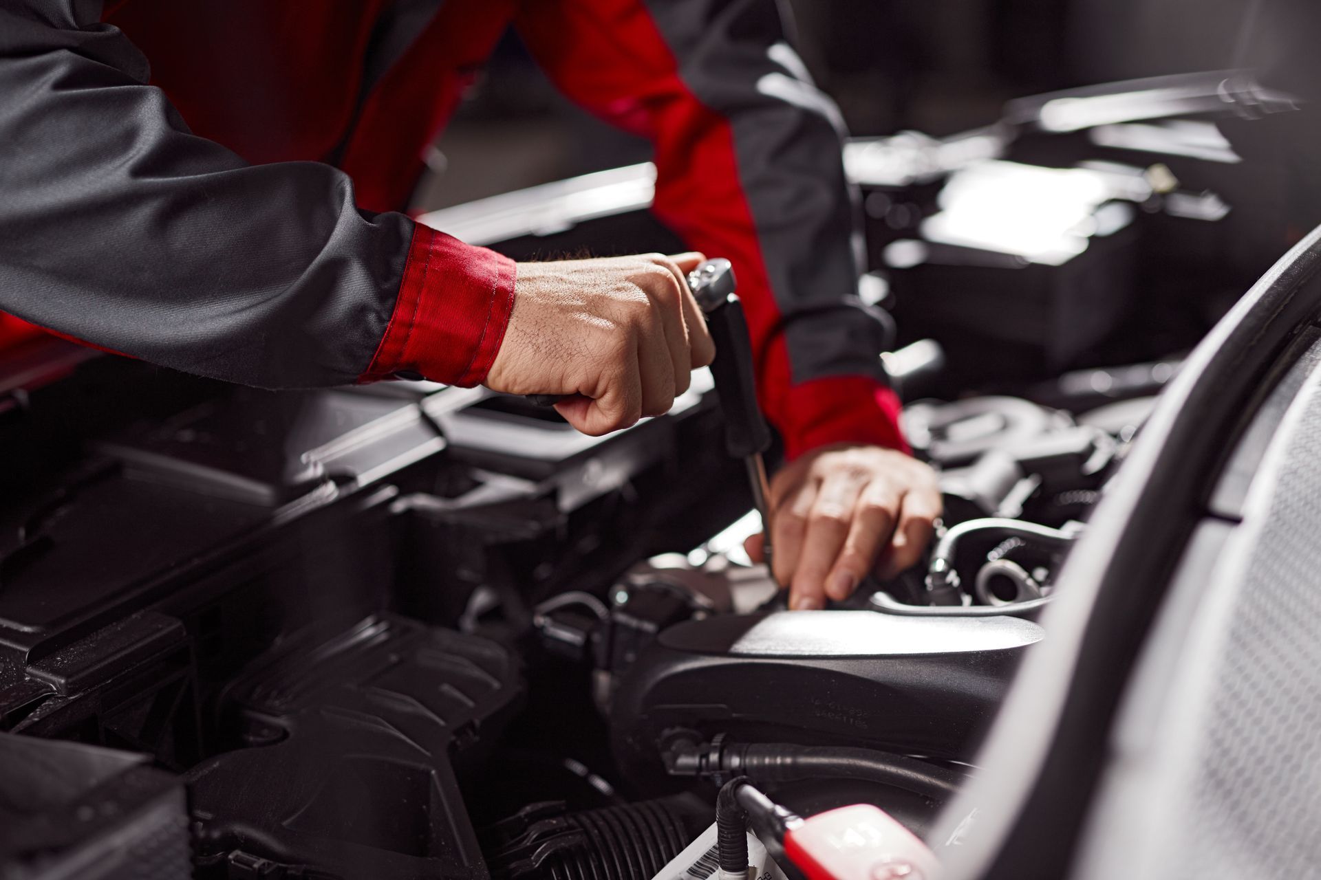 A mechanic in a red and grey uniform uses a screwdriver to work on a car engine.