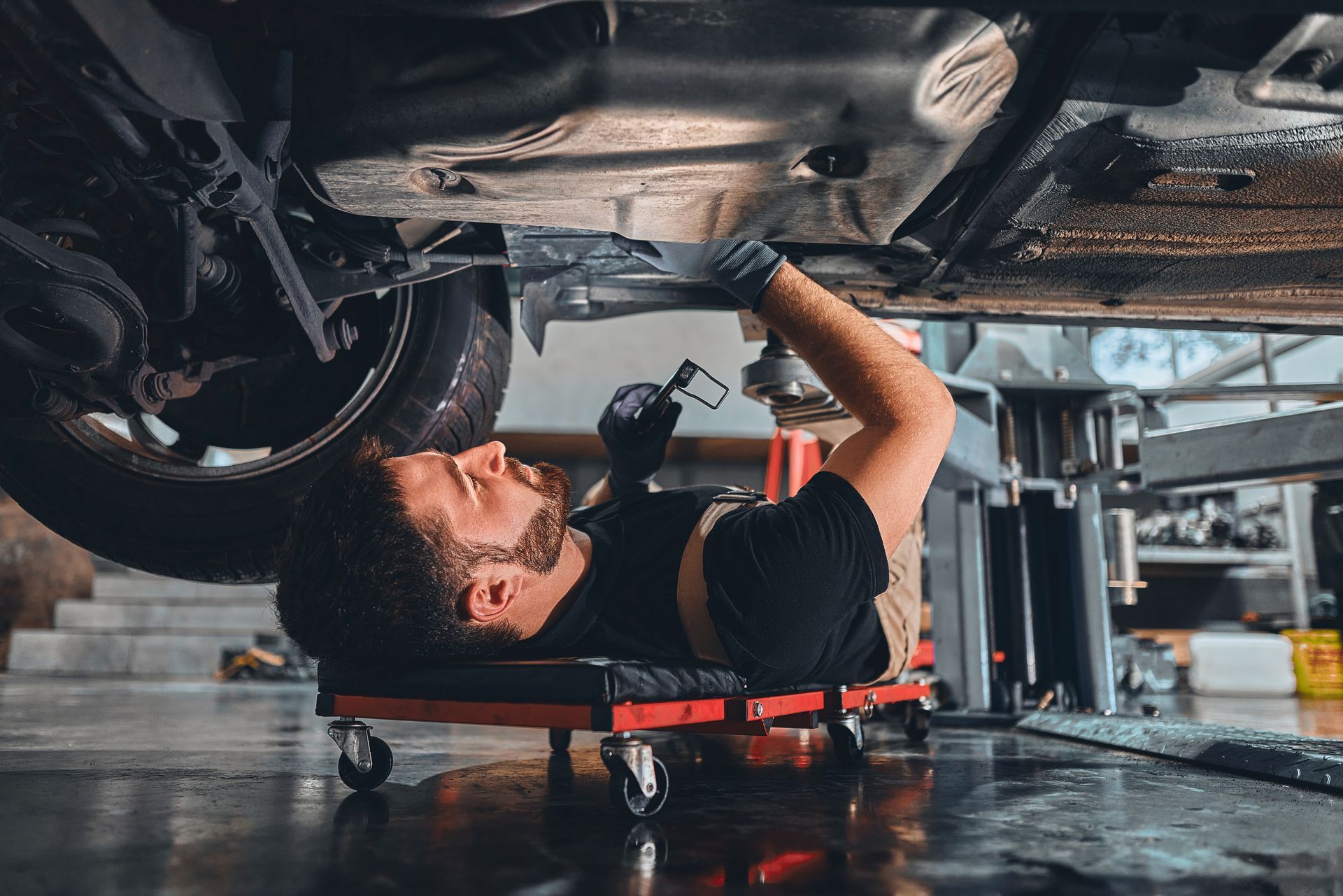 A mechanic in black work clothes lies on a creeper, inspecting the undercarriage of a car in a garage.