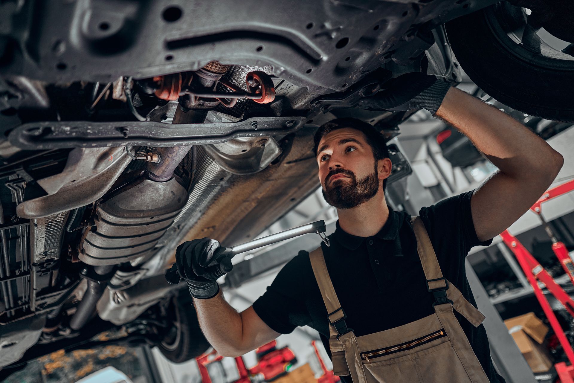 A mechanic in a black shirt and beige overalls repairs the undercarriage of a car in a workshop.