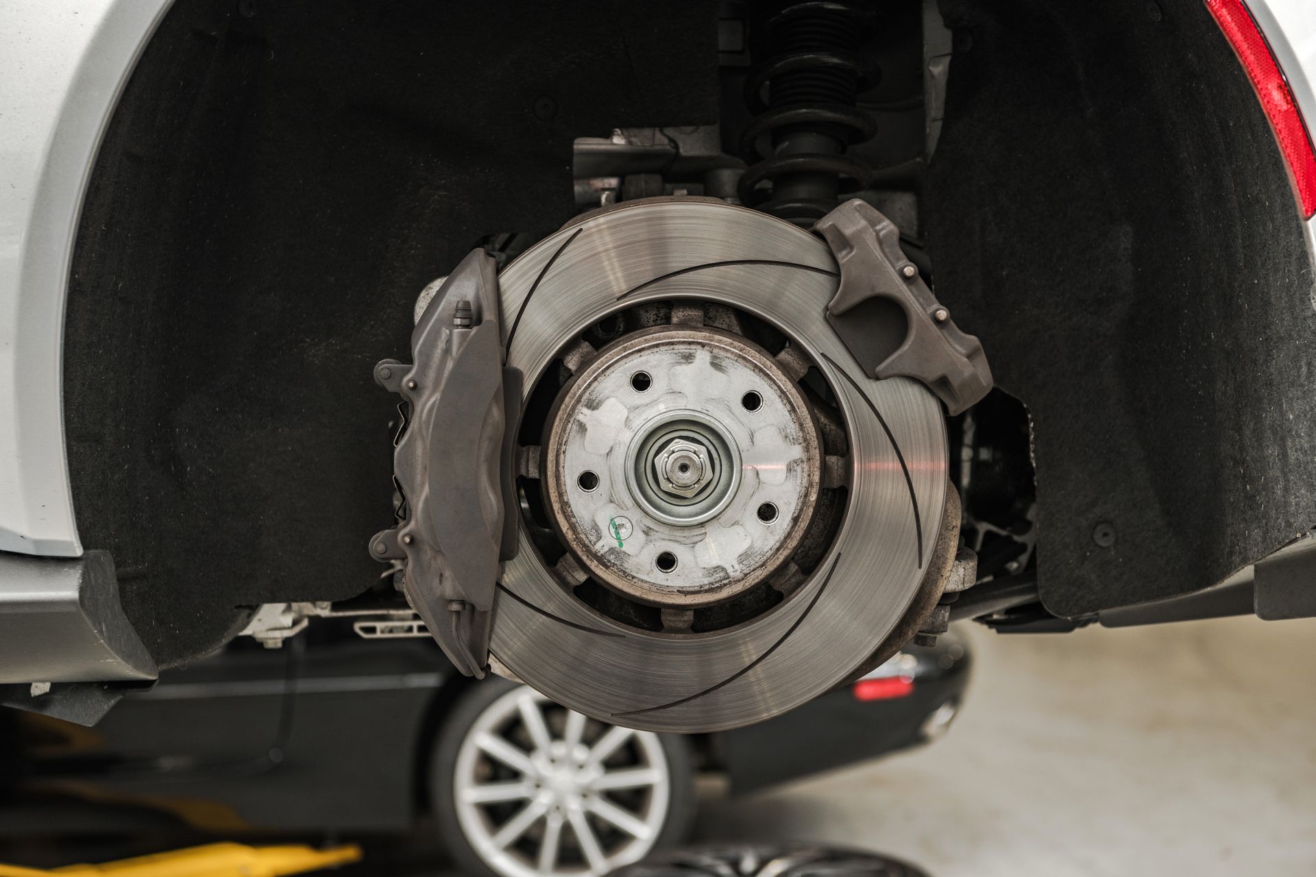 A closeup of a car's exposed wheel hub assembly with a slotted brake rotor and caliper, viewed in an auto repair shop.