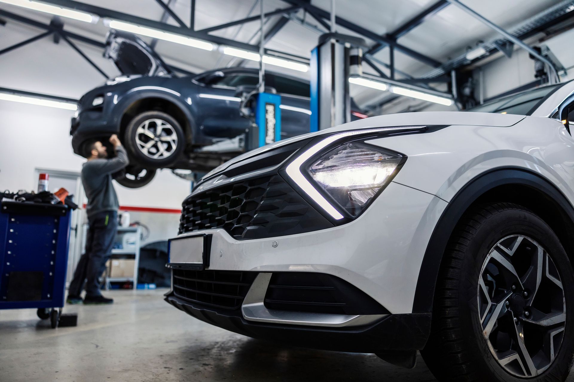 A mechanic works on a blue car raised on a lift in a workshop, with a white SUV in the foreground.