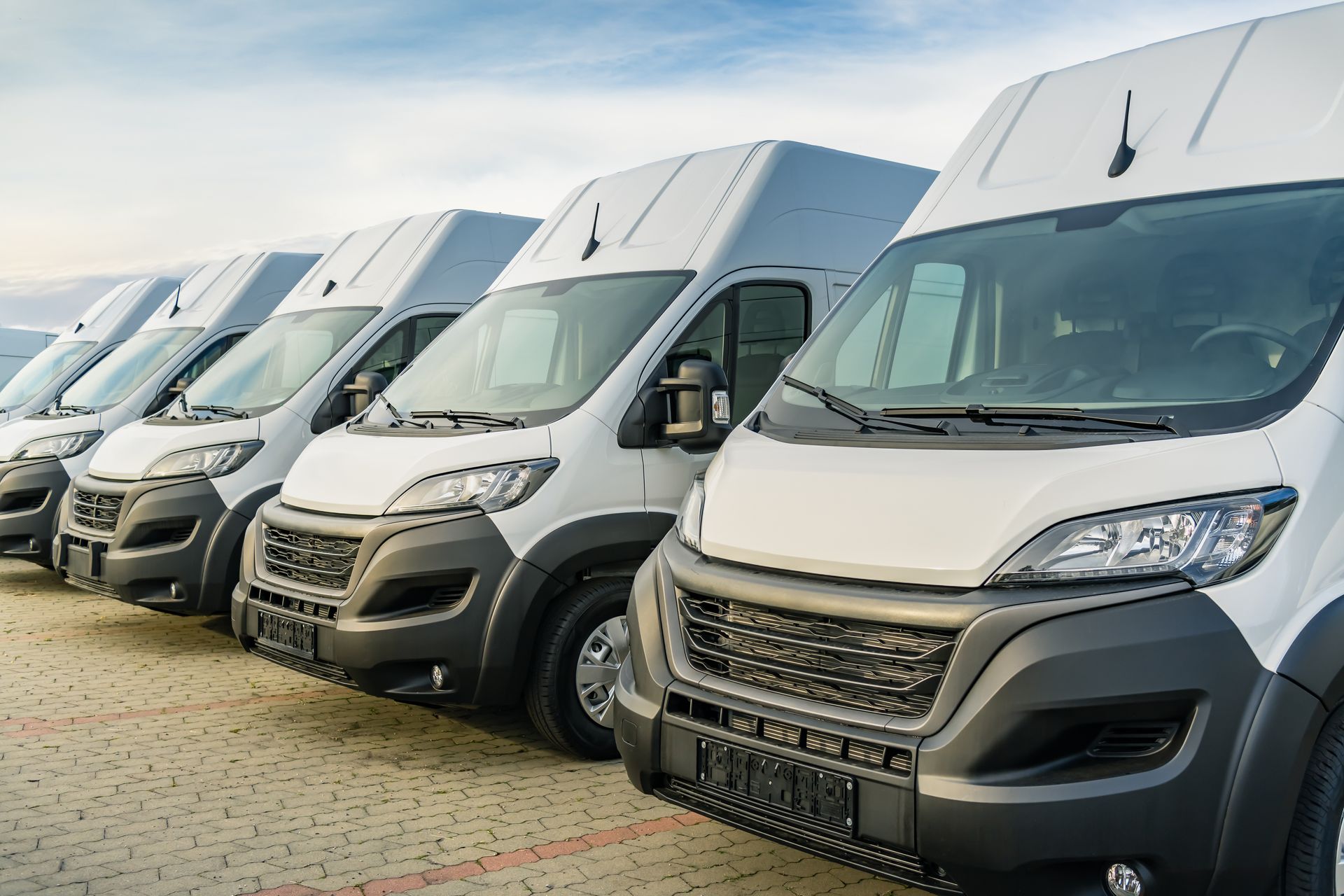 A row of white cargo vans parked on a paved lot under a cloudy sky.