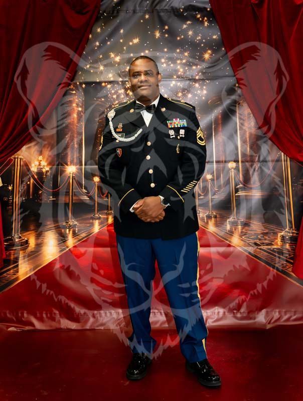 A person in a U.S. Army dress uniform posing on a red carpet in front of red curtains and a festive, starry backdrop.