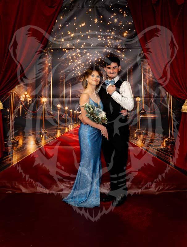 A couple poses for a formal portrait on a red carpet in a room decorated with string lights and draped red curtains.