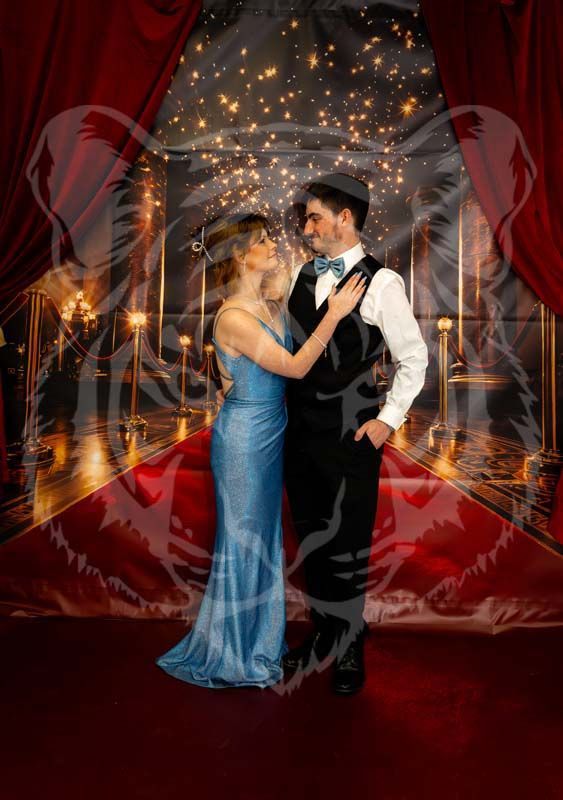 A couple dressed for a formal event stands together on a red carpet in front of a backdrop with lights and curtains.