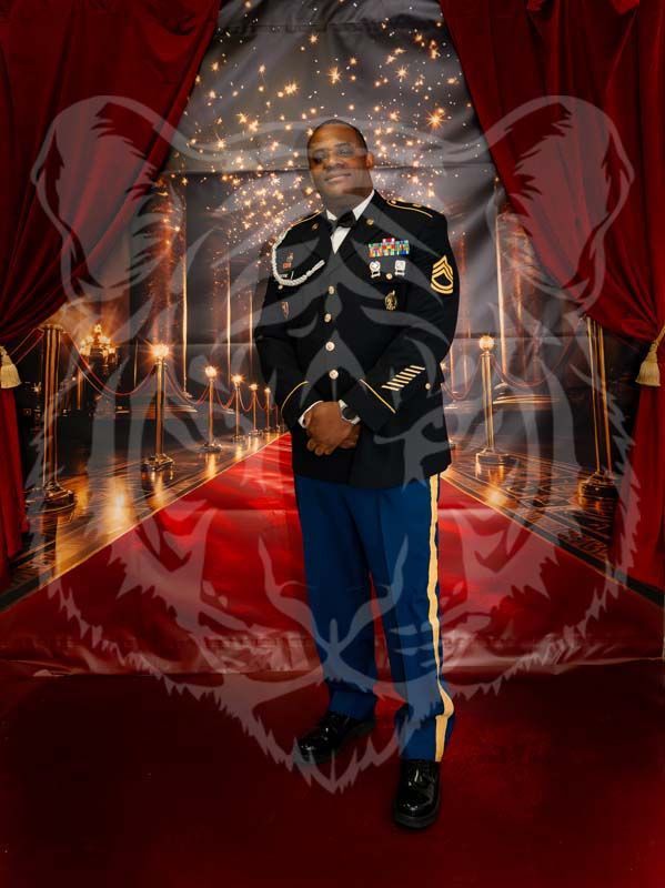 A person in a U.S. Army dress blue uniform stands on a red carpet in front of a backdrop resembling a decorated hallway.