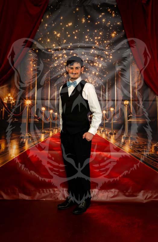 A person wearing a suit, vest, and light blue bowtie stands on a red carpet in front of a theater-themed backdrop.