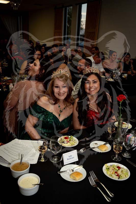 Two people wearing tiaras sit at a banquet table with salad and bread in a dimly lit, festive setting.