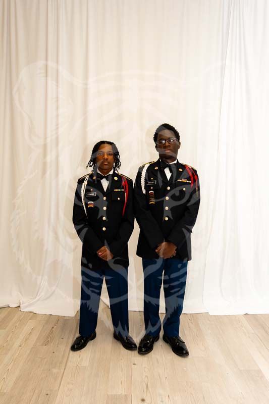Two individuals in formal military-style dress uniforms stand side-by-side against a white curtain backdrop.