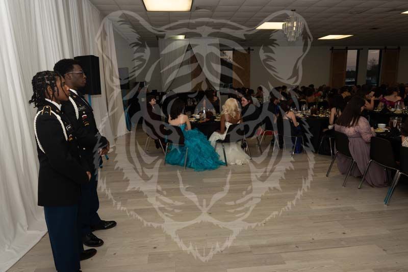 Two military personnel in dress uniforms stand at attention in a banquet hall, observing a crowd seated at tables.