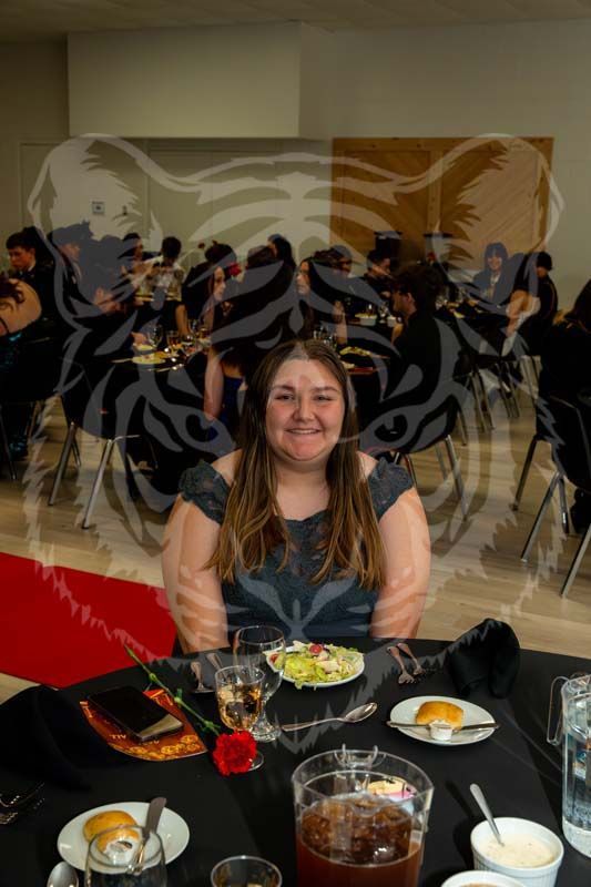 A person smiles at a table during a banquet, with place settings, food, and a red rose visible in the foreground.