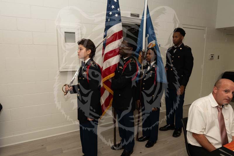 A color guard in uniform carries two flags in a room, with a person seated in the foreground.