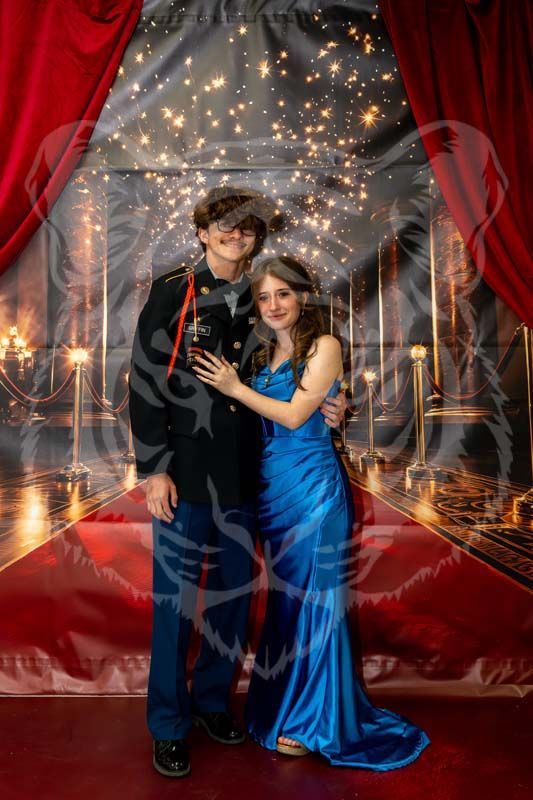 A couple stands posed in formal wear before a backdrop featuring red curtains, stanchions, and a starry, bokeh background.