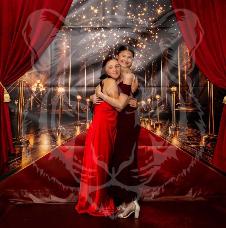 Two people embrace while wearing formal dresses in front of a red carpet backdrop with red curtains and sparkling lights.