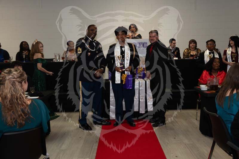 Two individuals in military uniforms stand on a red carpet during an indoor awards ceremony with guests seated nearby.