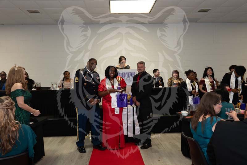 People in formal attire gather at a banquet, with three individuals standing on a red carpet in front of a stage.