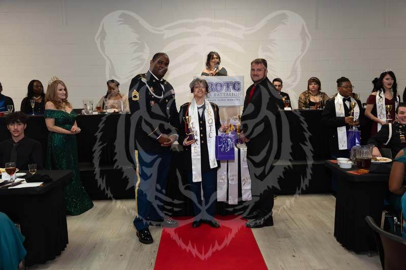 Three individuals stand on a red carpet in front of a tiger mural at an event, flanked by tables with seated guests.