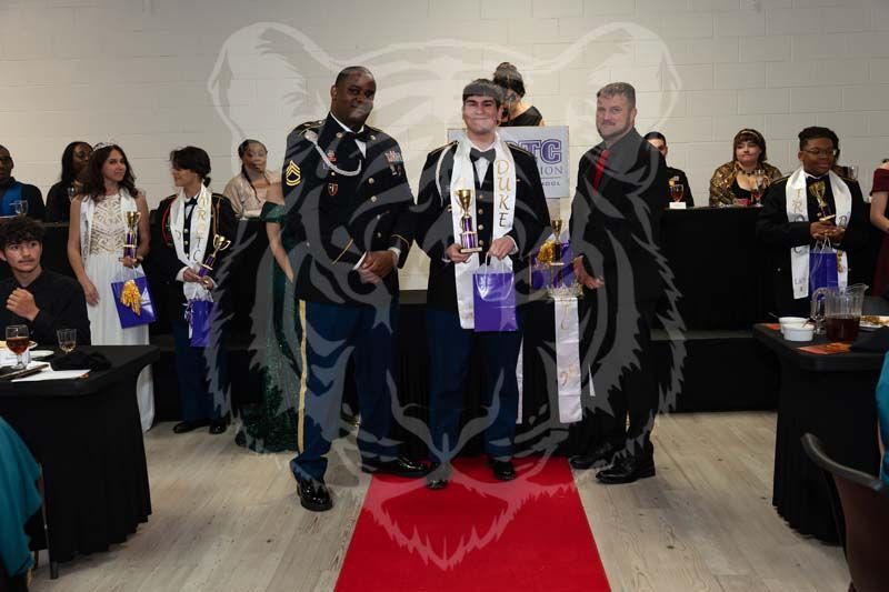 Three individuals in formal wear and military attire stand on a red carpet at an awards banquet held in a hall.