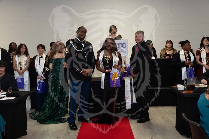 A group stands on a stage for an awards ceremony, with two central figures holding trophies on a red carpet.