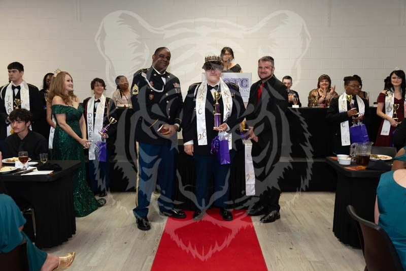 Formal event with people in evening attire standing on a red carpet, featuring two central awardees wearing sashes.