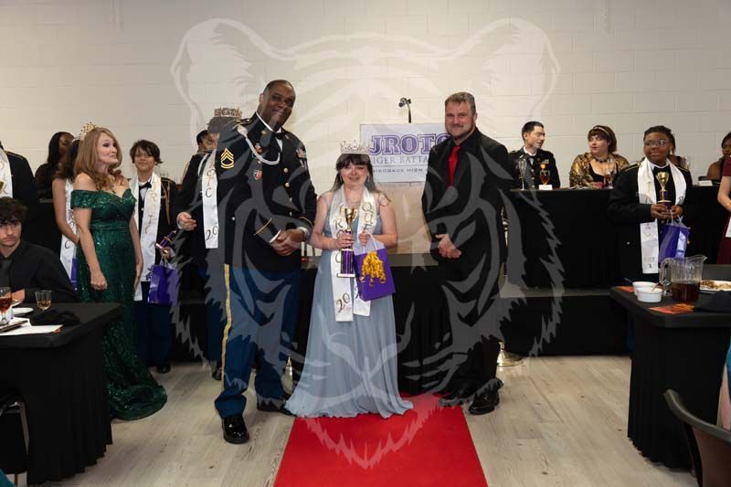 People standing on a red carpet at a formal event, including a person in a crown and light blue gown, inside a building.