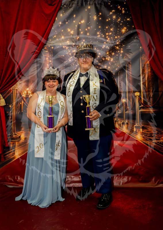 A pair in formal wear, wearing sashes and holding trophies, stands on a red carpet in front of a celebratory backdrop.